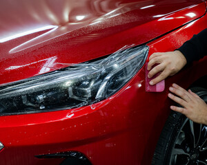 The master applies vinyl film to the headlight of a red car. Closeup view on worker detailer hand smoothing with a scraper protective film. 