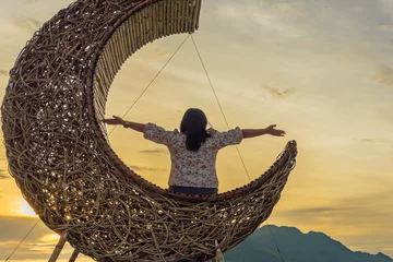 Tuinposter Woman sit on crescent moon chair made of rattan for relaxation on bridge in paddy field with beautiful scenic in evening. Decorative wooden moon furniture as sitting chair for viewpoint in rice field © JinnaritT