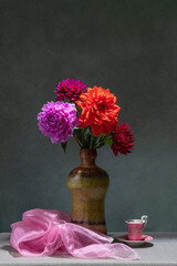 Still life with flowers. Pink, red dahlias in a dark vase, pink fabric, small pink cup, dark background