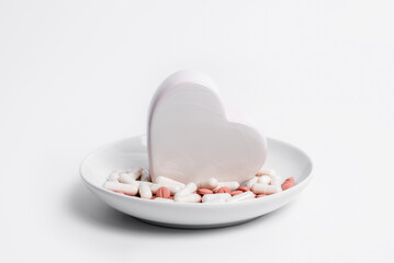 Pills and ceramic heart on white plate. Concept of cardiac diet and cardiology treatment of cardiovascular diseases with medication, dietary supplements and vitamins for heart health