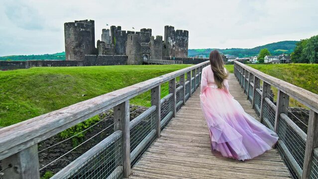 A beautiful woman in a princess dress visits Caerphilly Castle in Wales. A tourist girl in a long dress enjoys a moated fortress with an imposing gatehouse, a fortified dam and a tower.