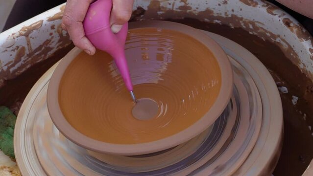 Close up real time shot of potter's  hands applying  glaze on a clay plate using potters circle.