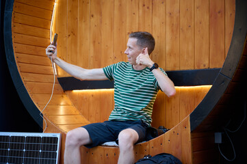 Excited photographer looks at the photos he just took. Young photographer taking a selfie. Handsome male sitting on round bench with solar panel on background.