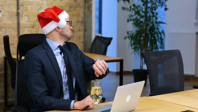 Caucasian man is working on his computer, and looking into his watch, he is taking off Santa's head, and drinking a glass of sparkling wine. Realtime video