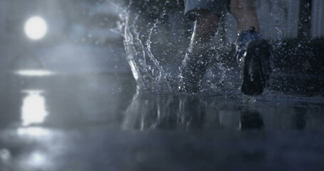 Child runs in water puddle with rain boots during rainy season in slow-motion with droplets flying...