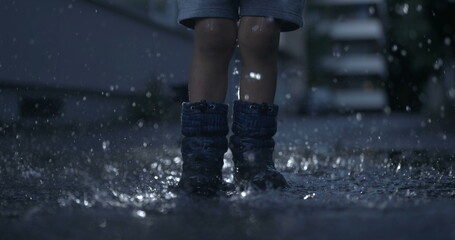 Excited child wearing rain boots jumps into water puddle in slow-motion capturing droplets in the...