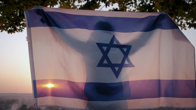 A woman stand with the flag of Israel against a clear sky at sunset.