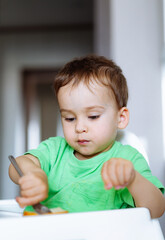 Curious Toddler Learning to Eat Independently. A little boy sitting in a high chair with a knife and fork in his hand