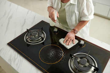 Senior woman cleaning greasy stove with detergent, view from above