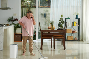 Cheerful elderly woman talking on phone with friend when wiping floor at home