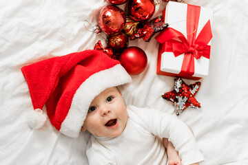 Christmas toddler in Santa hat lying on bed with gift box and festive baubles top view