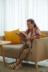 Senior woman sitting on couch in living room and reading book