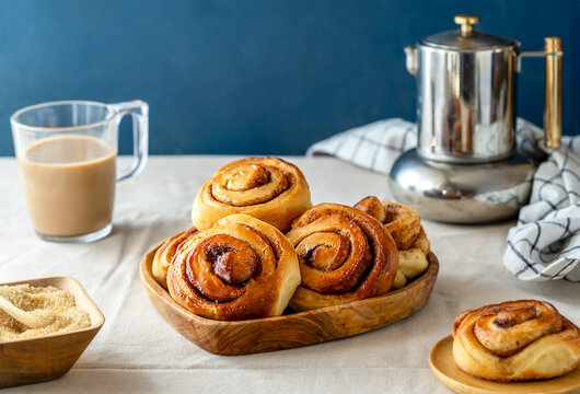 Homemade fresh baked cinnamon rolls in a wooden tray, cinnamon sticks, sugar, coffee-pot, napkin and cappuccino 