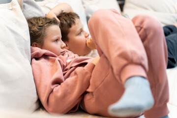 Brother and sister lying comfotable sofa and waching TV exciting movie. Boy biting his nails. Common interests among children. Ordinary everyday life of a simple family. 