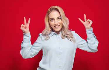 Playful woman gesturing peace sign with fingers, isolated on red background. Cheerful girl gesturing V sign