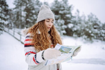 Curly-haired young woman standing in the wild with a map and compass in a snowy forest. Winter is approaching, the first snowfall. Adventure concept. - Powered by Adobe