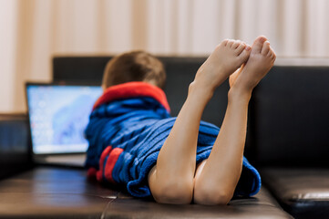 A boy child in pajamas lies on a black leather sofa in the evening, watching a movie on a laptop...