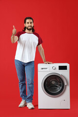 Young man near washing machine showing thumb-up on red background