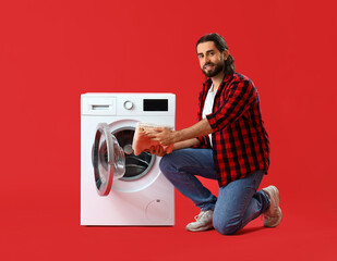 Young man putting laundry into washing machine on red background