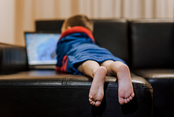 A small boy child in pajamas lies on a black leather sofa in the evening, watching a movie on a...