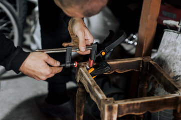 An adult male professional woodworker carpenter glues a chair, repairs, restores furniture indoors, workshop, clamping wooden products with a clamp. Photography, work concept, portrait.
