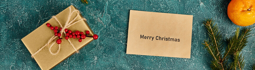 Merry Christmas envelope near mandarin and gift box with holly berries on blue texture, banner