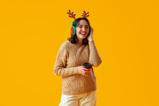 Young woman in reindeer horns with cup of coffee on yellow background