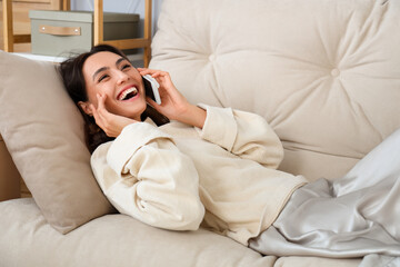 Young woman in warm sweater talking by mobile phone on sofa at home