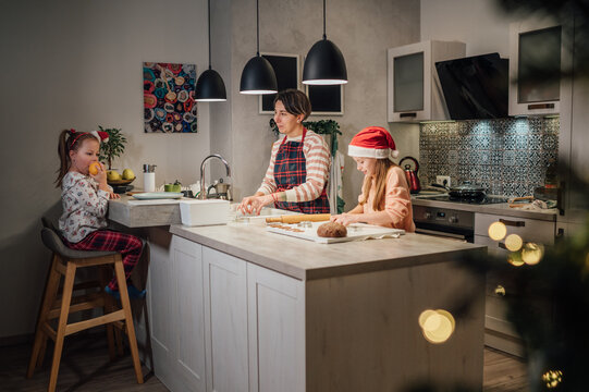 Cute little girls in red Santa hat with mother making homemade Christmas gingerbread cookies using cookie cutters together in home kitchen. Happy family holidays preparation and childhood concept.