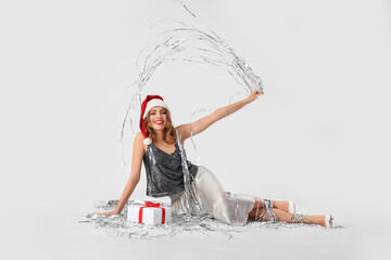 Beautiful young woman in Santa hat with gift and silver tinsel on light background