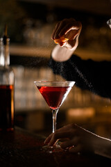 Female hand squeezes orange slice and sprinkles orange peel on cocktail in cocktail glass