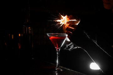 Transparent cocktail glass with brown alcoholic drink and lit match above it stands on bar counter