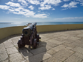 Canhão de ferro junto a um muro apontado na direção do mar no horizonte num dia de sol. Canhão medieval em cima de suporte com rodas de ferro. - obrazy, fototapety, plakaty