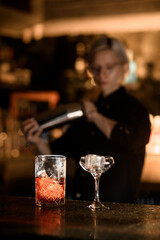 Glass for mixing a drink and a stemmed glass with ice are on the bar counter on the background of a female bartender