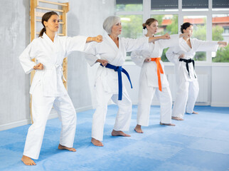 Focused beginner young female karateka in kimono standing in attacking stance, practicing punches or hand striking techniques as part of kata with group of women