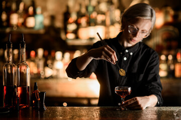 Female bartender in glasses adding a green leaf to a brown cocktail in a stemmed glass