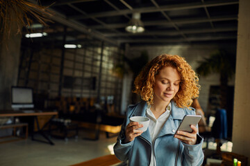 Young woman holding smartphone and coffee mug in office