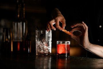 Female hands roll an orange peel into a tube over a glass with a cocktail