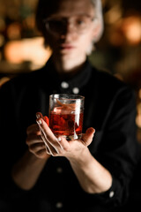 Female bartender shows in her palm a glass filled with an iced cocktail and a piece of orange peel