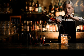 Female bartender pours an alcoholic drink into a mixing glass from a jigger and a bottle with a geyser - obrazy, fototapety, plakaty