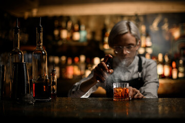 Female bartender squatting at the level of a glass sprays a ready-made cocktail in a glass - obrazy, fototapety, plakaty