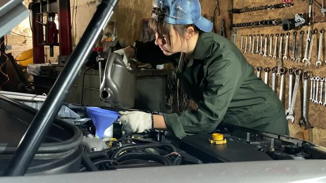 Young woman in coveralls works at car service station conducts an oil change. Female mechanic pouring genuine engine oil with a funnel into the oil filler neck indoors. Automotive tools on background