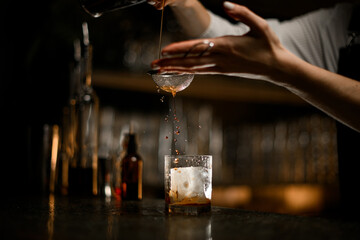 Female hands pours a cocktail through a strainer from a metal tumbler into a glass tumbler close-up