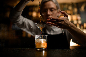 In the foreground is a glass with a piece of ice into which a cocktail is poured through a strainer on bar counter