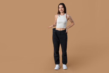 Fototapeta na wymiar Beautiful young sporty woman in loose jeans on brown background. Weight loss concept