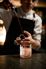 Female bartender simultaneously pours alcoholic drinks from a jigger and geyser bottle into frosted glass