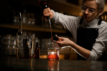 Female bartender mixes alcoholic drinks in a glass, pouring them simultaneously from a jigger and a...