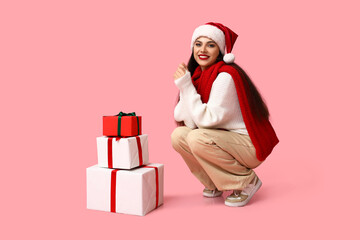 Beautiful young woman in Santa hat with Christmas gifts on pink background