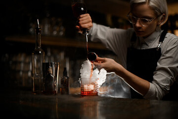 Female bartender mixes alcoholic drinks in a glass, pouring them simultaneously from a jigger and a geyser bottle with smoke