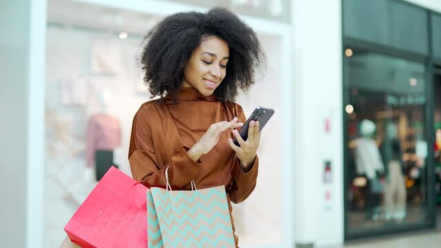Young beautiful happy woman shopper joyful customer stand in shopping center mall and uses a mobile phone browsing chatting typing smartphone Satisfied glad with gift color package bag in store